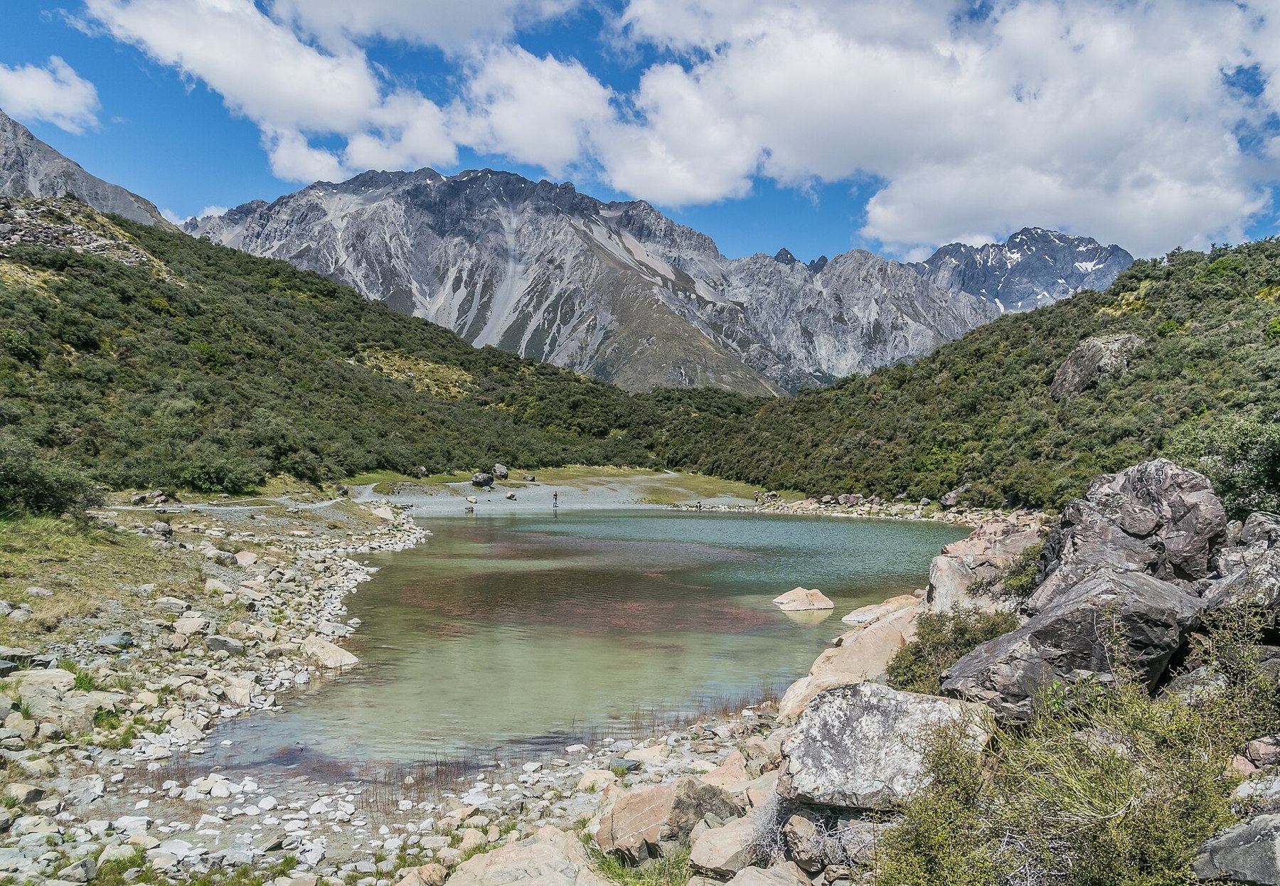 The rocky shore of a small lake, green shrubs and mountains beneath clouds