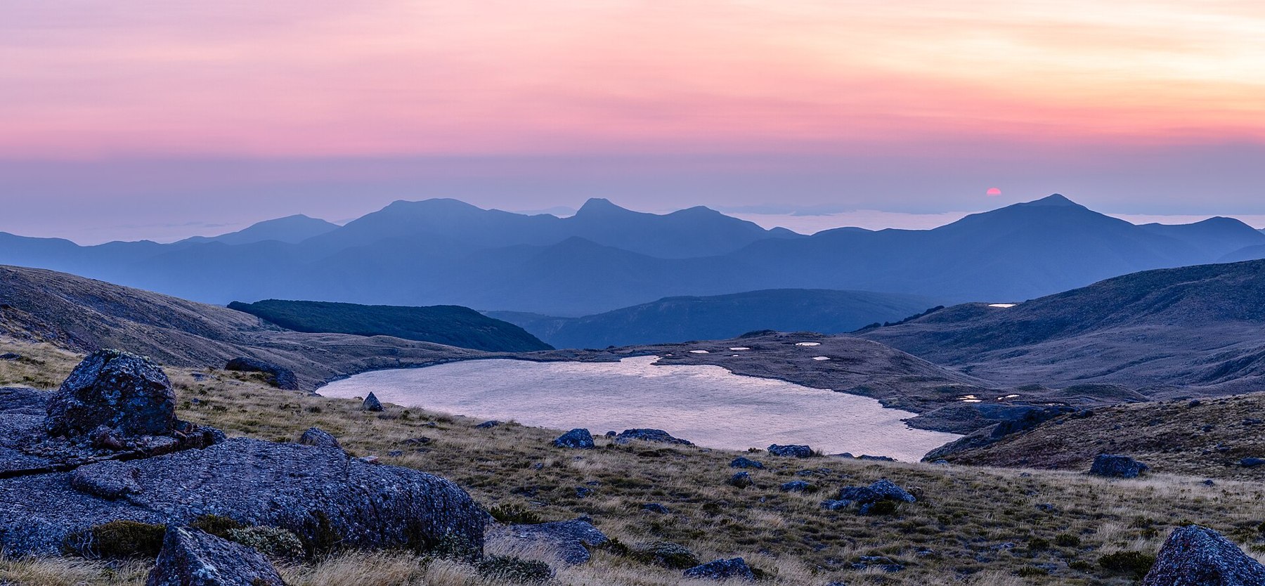A pink sky above blue mountain silhouettes and a lake among rocks